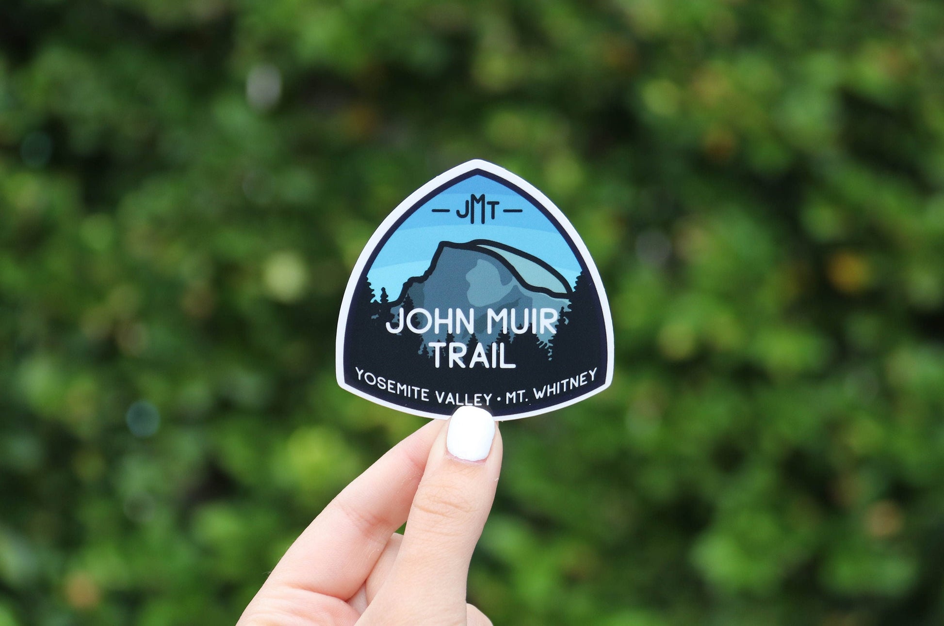 Two fingers hold the John Muir Trail sticker at arm's length, hedge blurred well behind it.