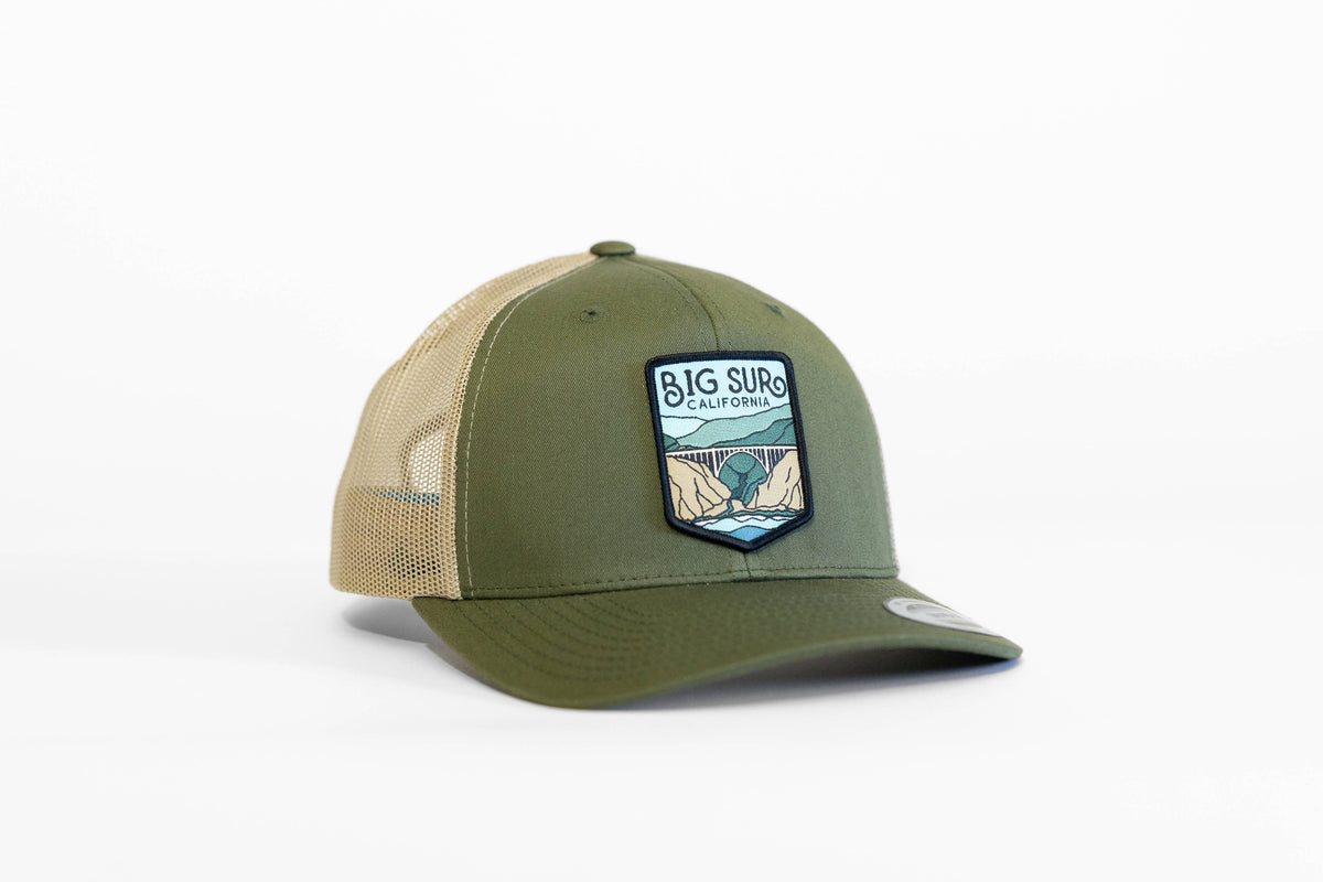 Angled on a white backdrop, the olive Big Sur California trucker hat shows its bridge patch and tan mesh.