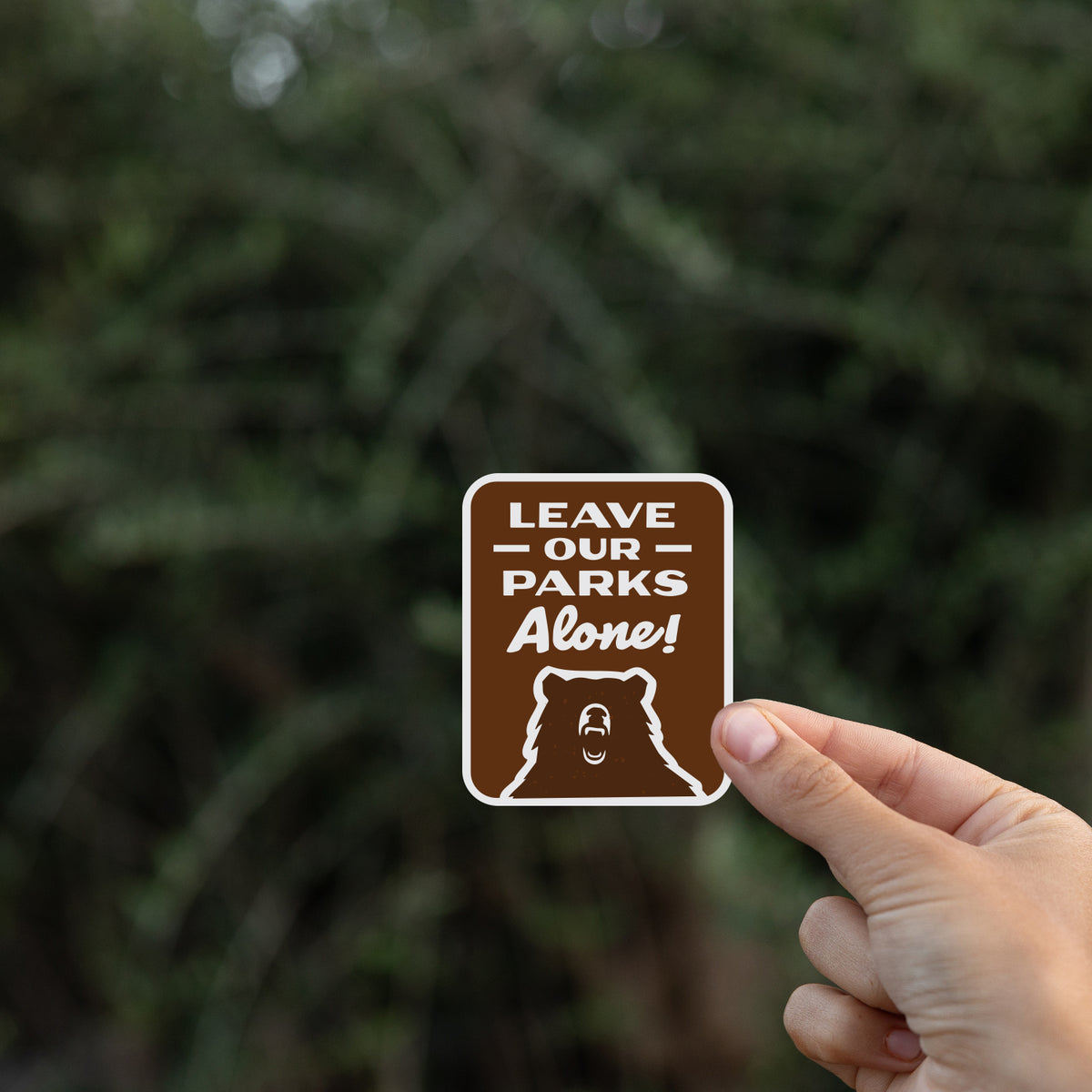 Pinched by a thumb, the Leave Our Parks Alone sticker hovers against a dark blurred hedge.