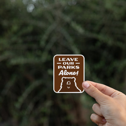 Pinched by a thumb, the Leave Our Parks Alone sticker hovers against a dark blurred hedge.