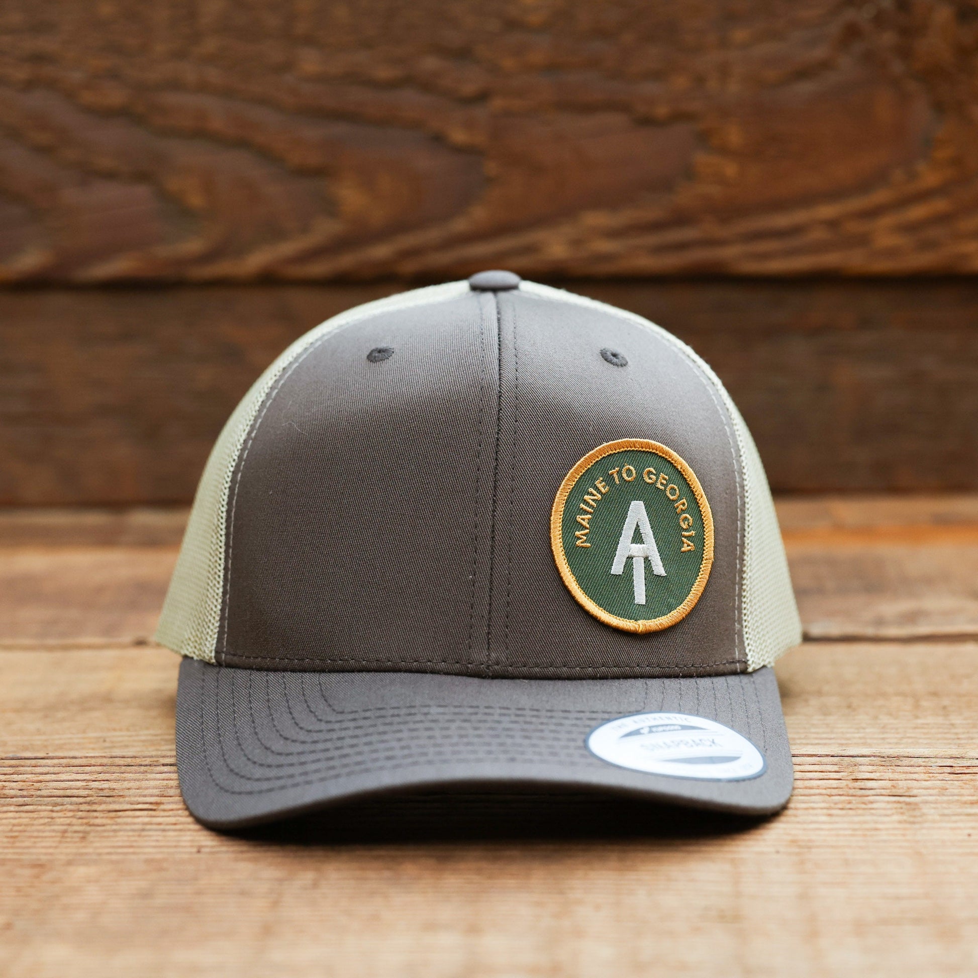 Centered on a plank wall, the brown Appalachian Trail trucker hat holds its green circular patch front and low.