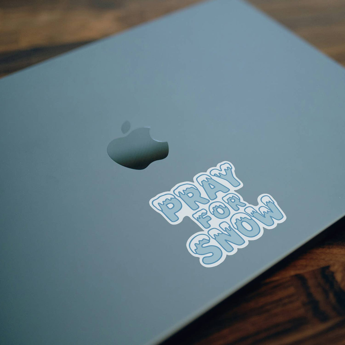 Stuck to a pale green laptop lid over dark wood, the Pray For Snow sticker shows its icy blue lettering.