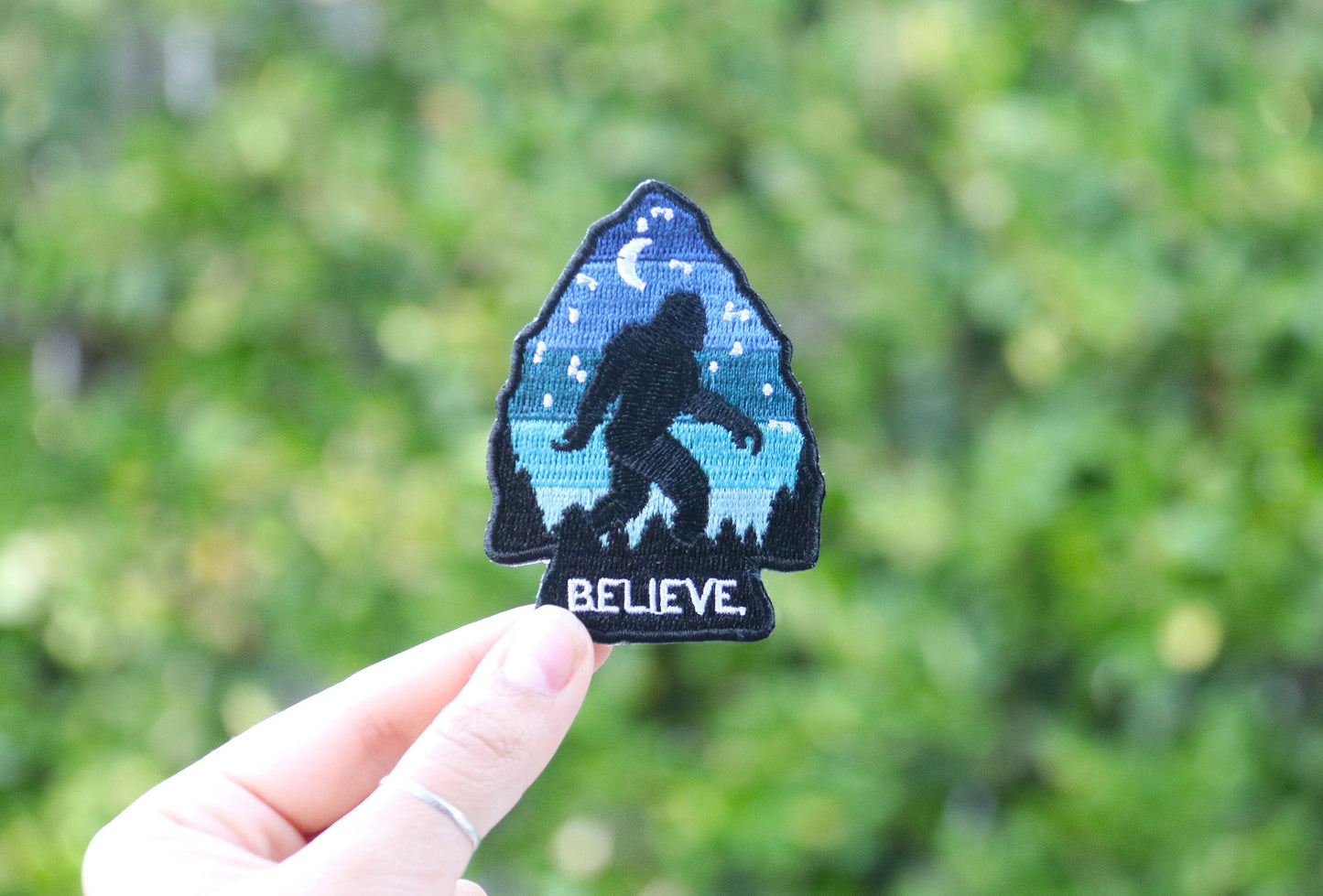 Held higher in daylight, the Bigfoot Believe patch catches sun with hedges soft behind the hand.