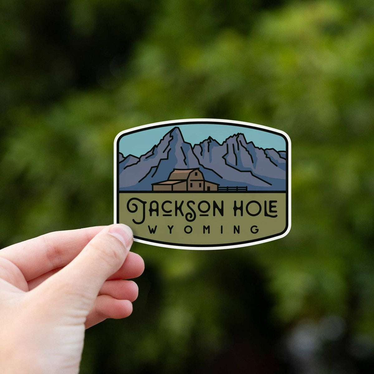 A hand grips the Jackson Hole Wyoming sticker from below with green summer foliage out of focus behind.