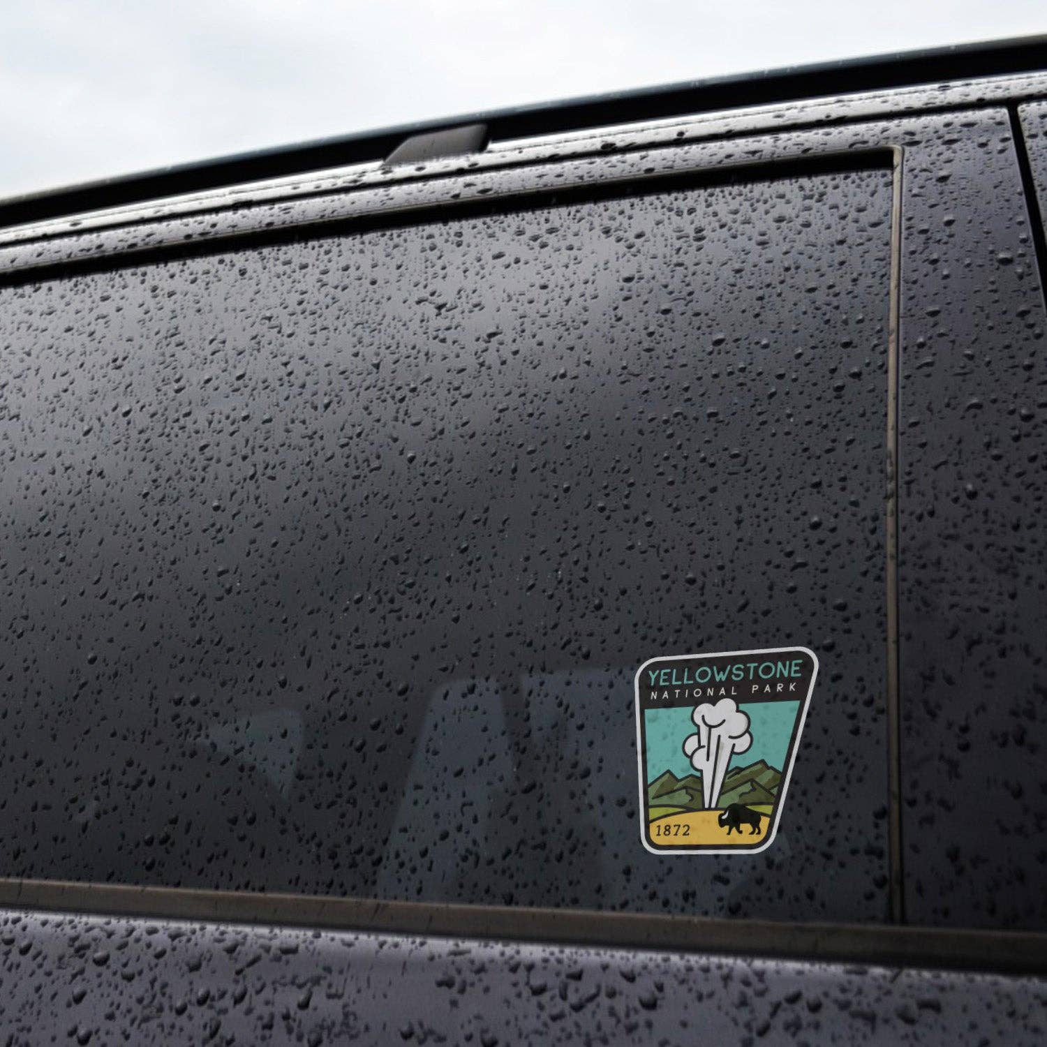 Rain beads across a black car window where the Yellowstone National Park sticker is applied near the frame.