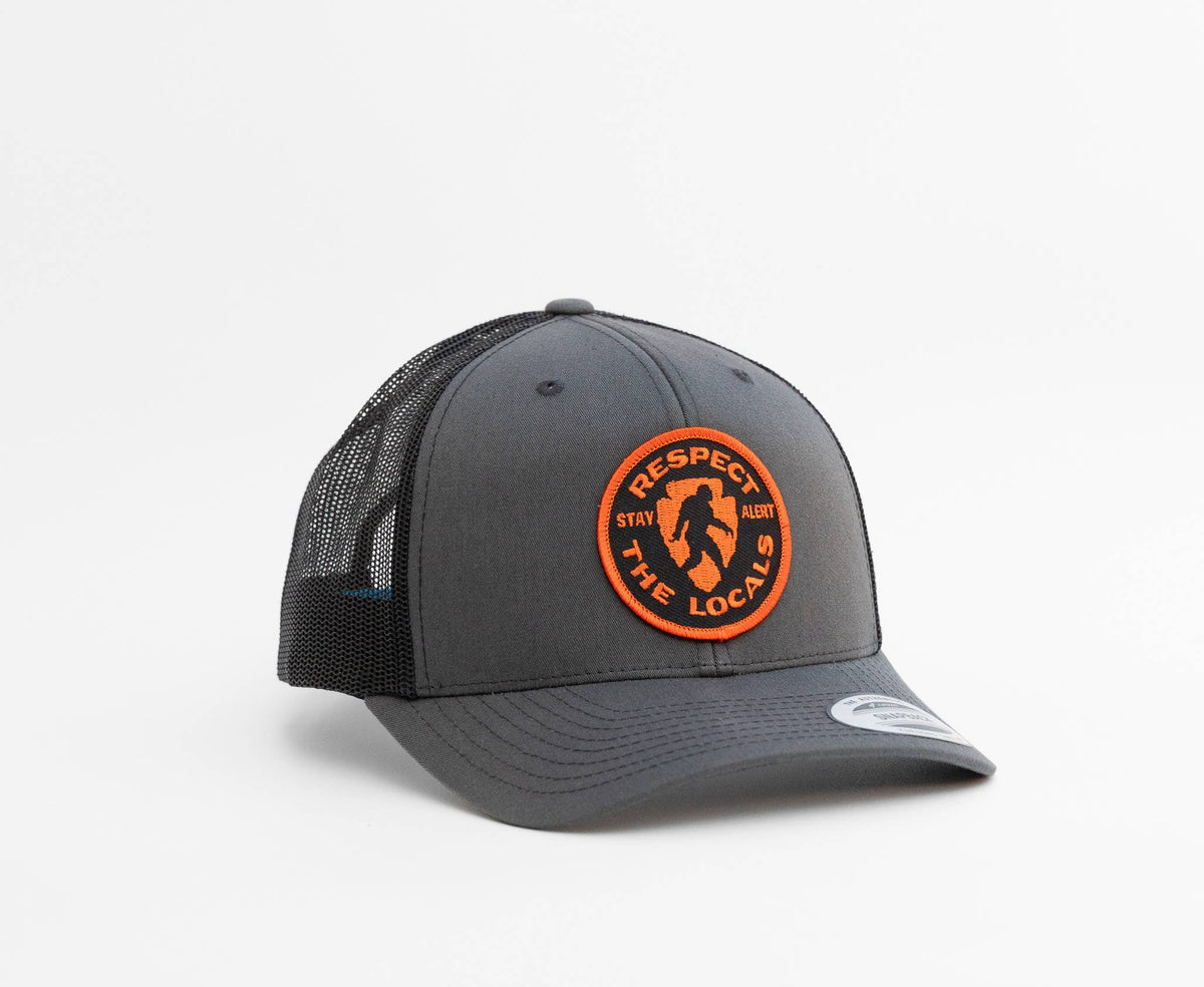 Charcoal and black Bigfoot Respect The Locals trucker hat turned three quarters, orange patch facing the camera.