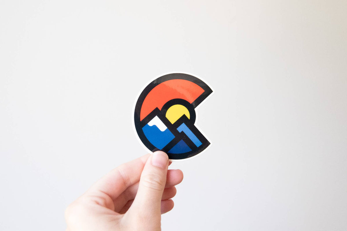 A hand holds the Colorado Mountains sticker against a plain white wall, its red C circling blue peaks and a yellow sun.