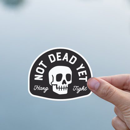 A thumb and finger hold the Not Dead Yet sticker against a soft grey expanse of still water.