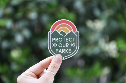 Farther back against blurred foliage, the Protect Our Parks sticker rests between finger and thumb of a ringed hand.