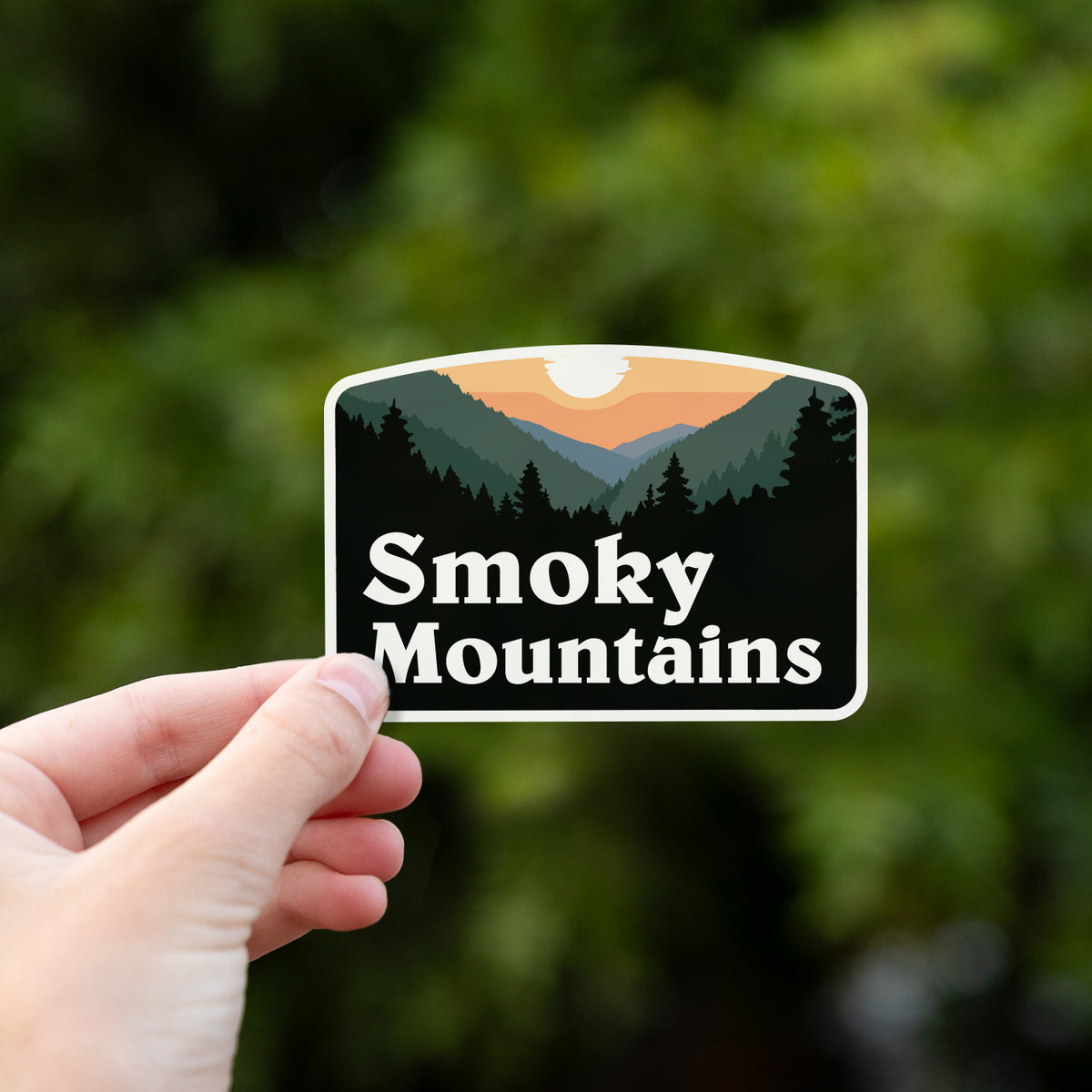 A hand holds the Smoky Mountains sticker up against a blurred wall of green hedge leaves.