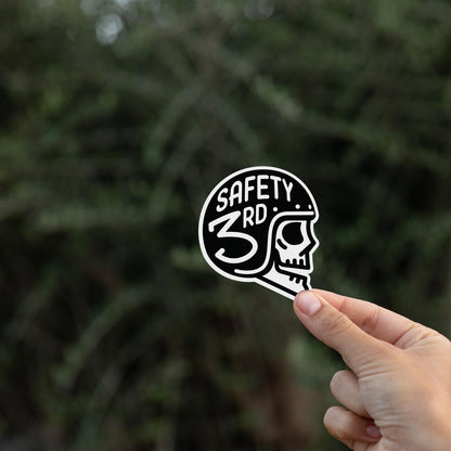 Held up against dark evergreen foliage, the Safety 3rd Helmet sticker shows its skull in profile.