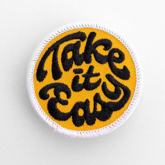 Yellow round Take it Easy embroidered patch with black script lettering and a white stitched border on grey.
