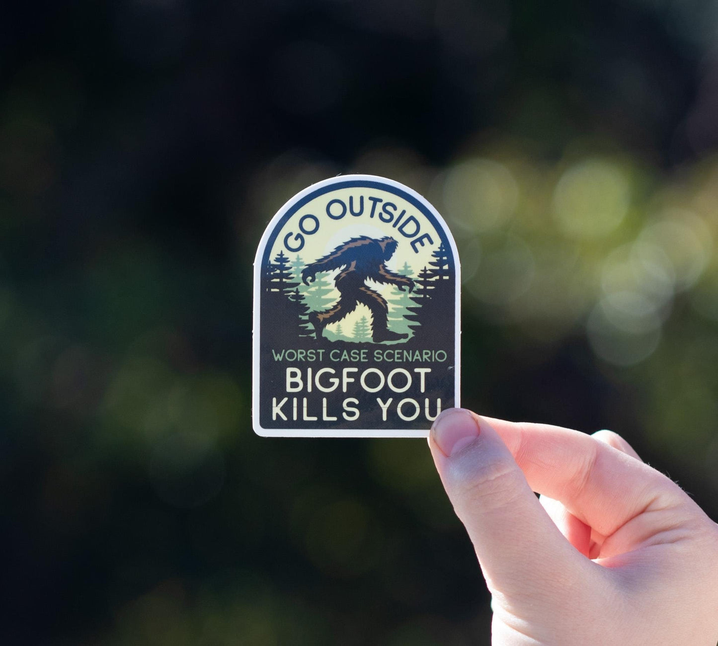 A hand holds the arched Go Outside Worst Case Scenario Bigfoot Kills You sticker against blurred dark green foliage.