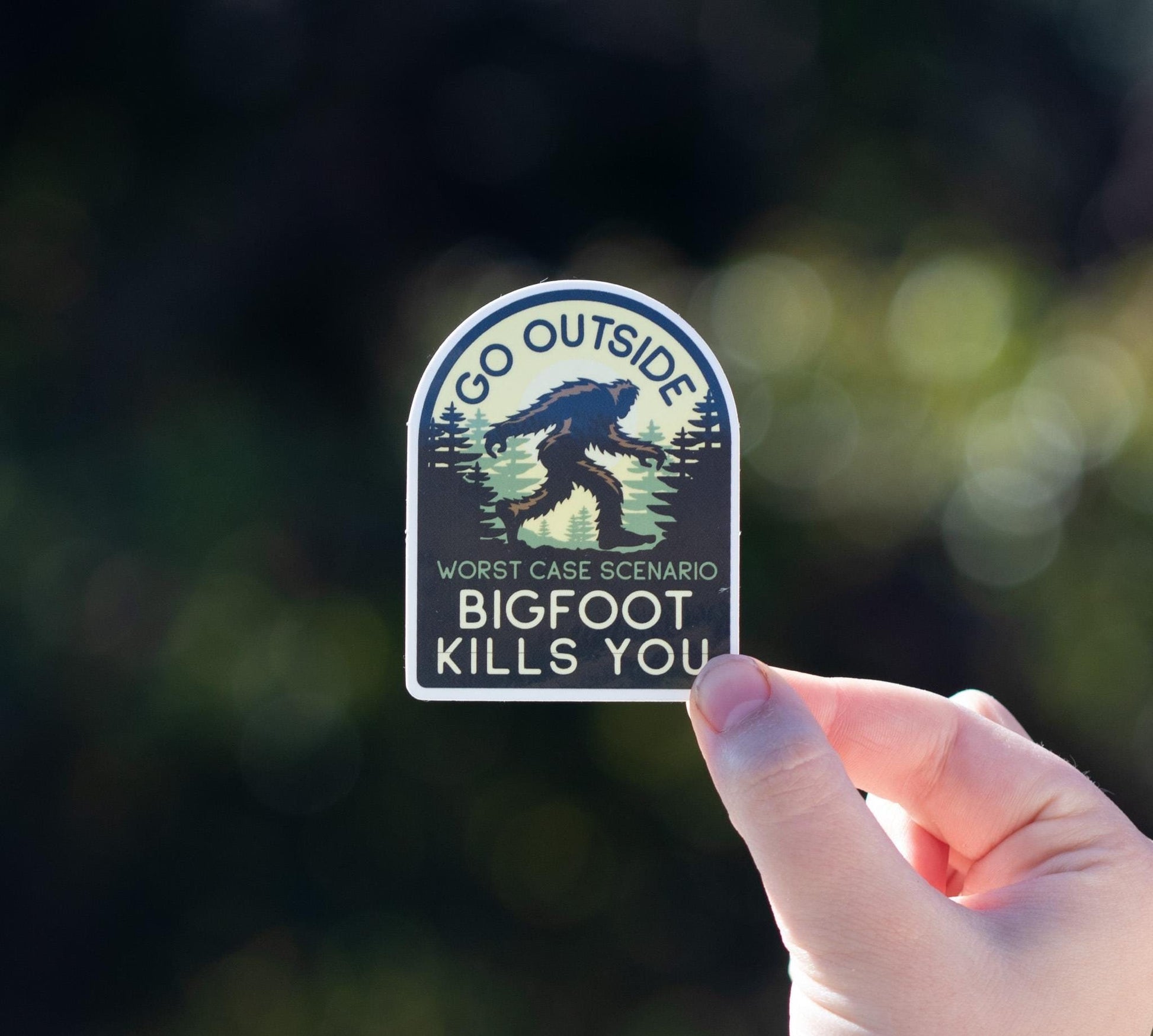 A hand holds the arched Go Outside Worst Case Scenario Bigfoot Kills You sticker against blurred dark green foliage.