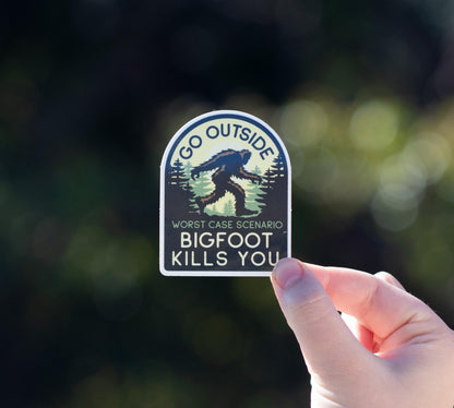 A hand holds the arched Go Outside Worst Case Scenario Bigfoot Kills You sticker against blurred dark green foliage.