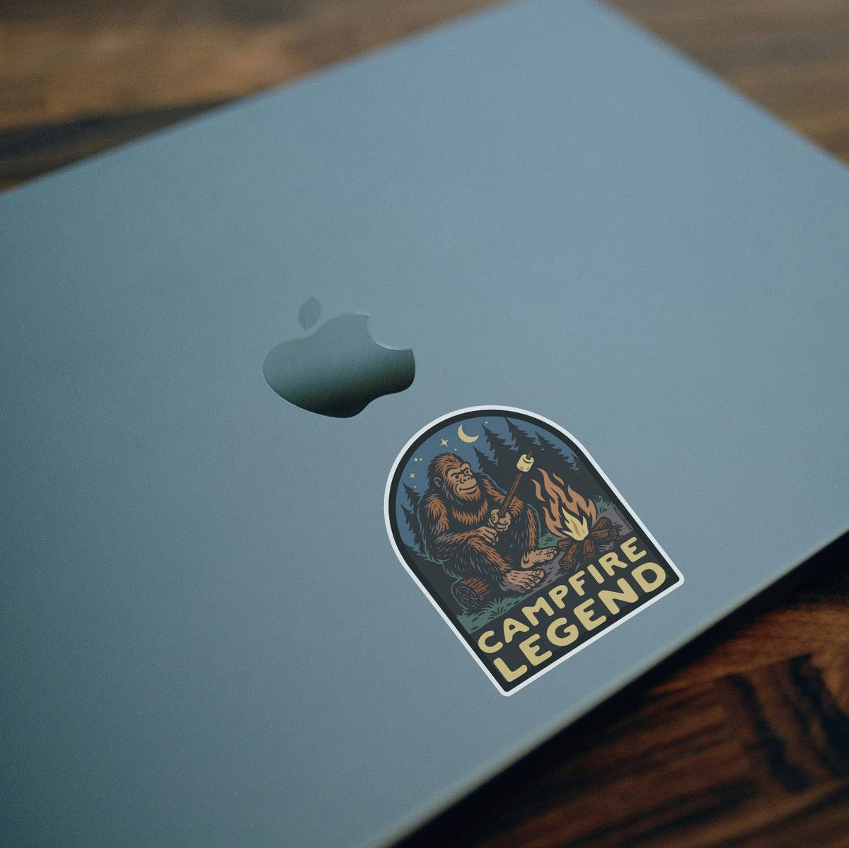 Stuck to a grey laptop lid on a wood table, the Campfire Legend sticker shows Bigfoot roasting a marshmallow.