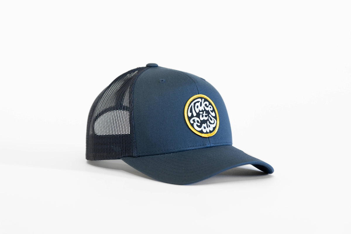Navy Take it Easy trucker hat turned three quarters, yellow ringed patch bright against the blue crown.