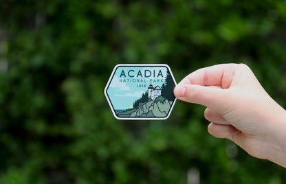 Held at arm's length before a leafy hedge, the small Acadia National Park sticker sits in the frame center.