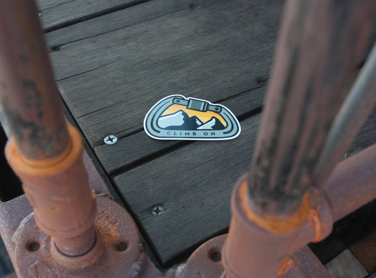 Stuck flat on dark deck planks between two rusted railing posts, the Climb On Carabiner sticker sits alone.