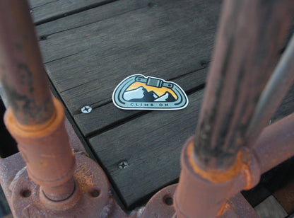 Stuck flat on dark deck planks between two rusted railing posts, the Climb On Carabiner sticker sits alone.
