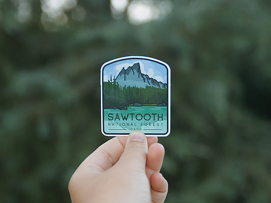 Backed by blurred evergreens, the Sawtooth National Forest sticker reads Idaho beneath the forest scene.