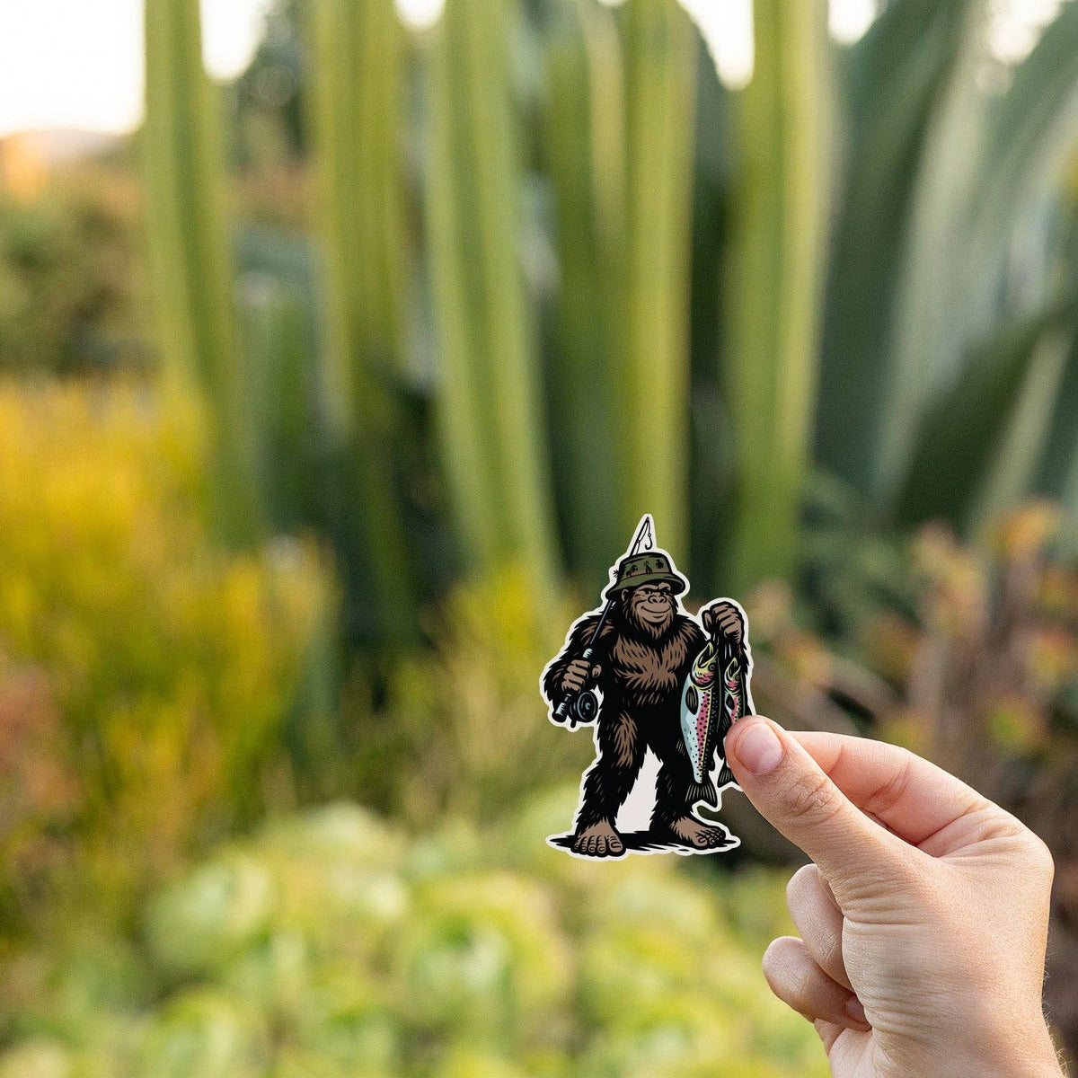 Tall cactus and succulents blur behind a hand holding the Bigfoot Fishing sticker at golden hour.