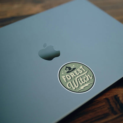 Stuck on the lid of a blue laptop beside the apple logo, the Forest Witch sticker sits on a wood desk.