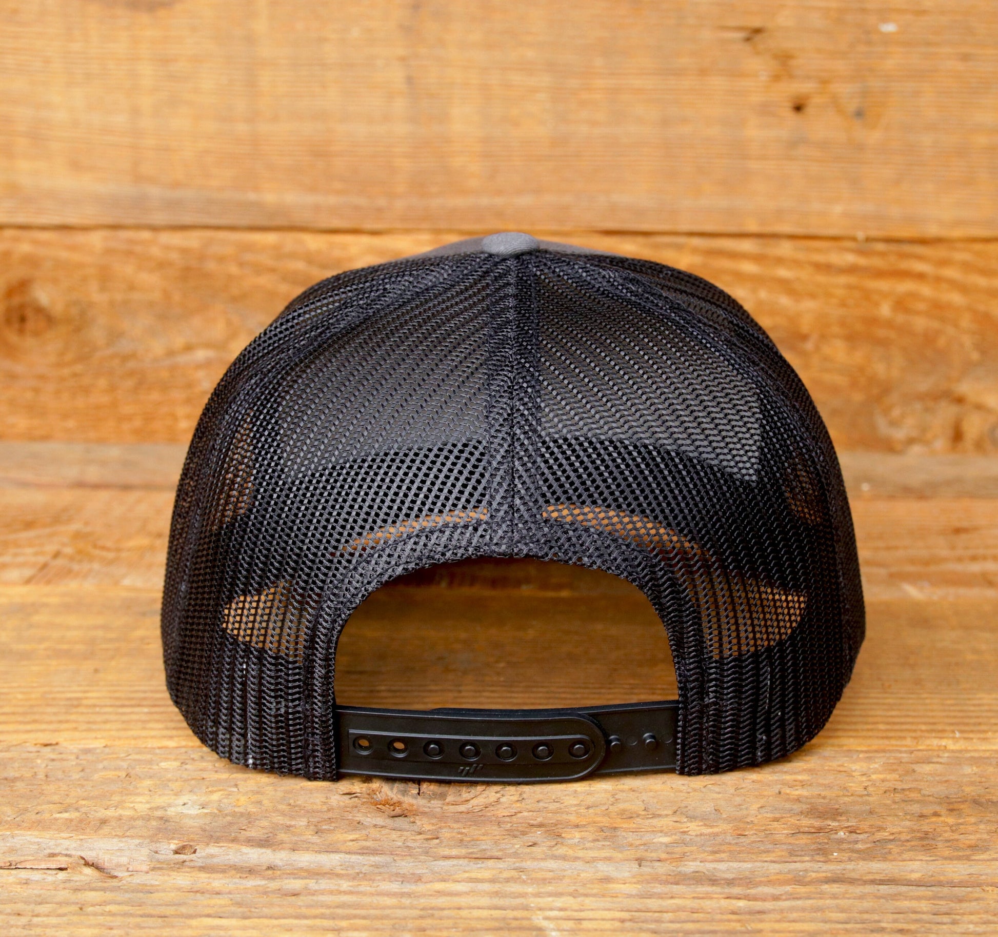 Against pale wood boards, the back of the Bigfoot Adventure trucker hat shows black mesh and snap closure.