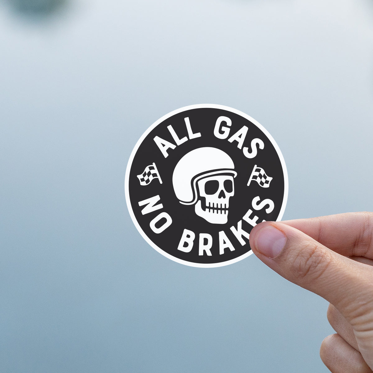 A thumb and finger hold the All Gas No Brakes sticker against a soft, pale blue blurred background.
