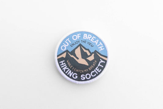 Flat on a white surface, the round Out of Breath Hiking Society patch shows its blue sky and mountain peaks.