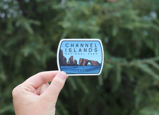 Held farther back against a blurred conifer, the Channel Islands National Park sticker shows blue water and rock spires.