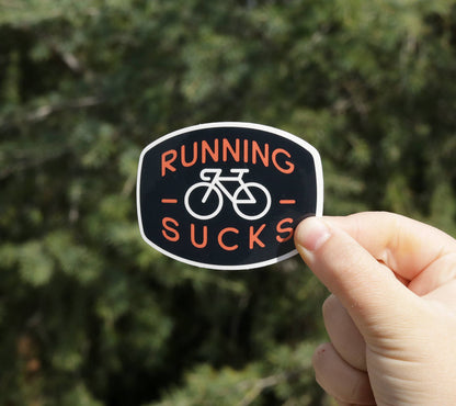 A hand pinches the Running Sucks sticker at arm's length before a blurred evergreen tree.