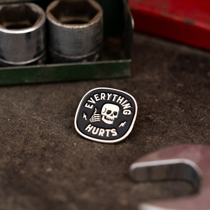 Resting on a grimy workbench near steel sockets, the Everything Hurts enamel pin sits beside a red toolbox.