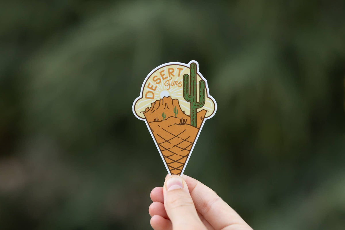 A hand holds up the Desert Time sticker, its cactus and butte cone shape bright against blurred green leaves.