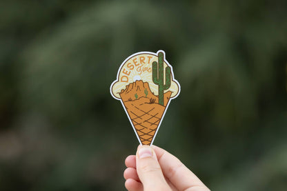 A hand holds up the Desert Time sticker, its cactus and butte cone shape bright against blurred green leaves.