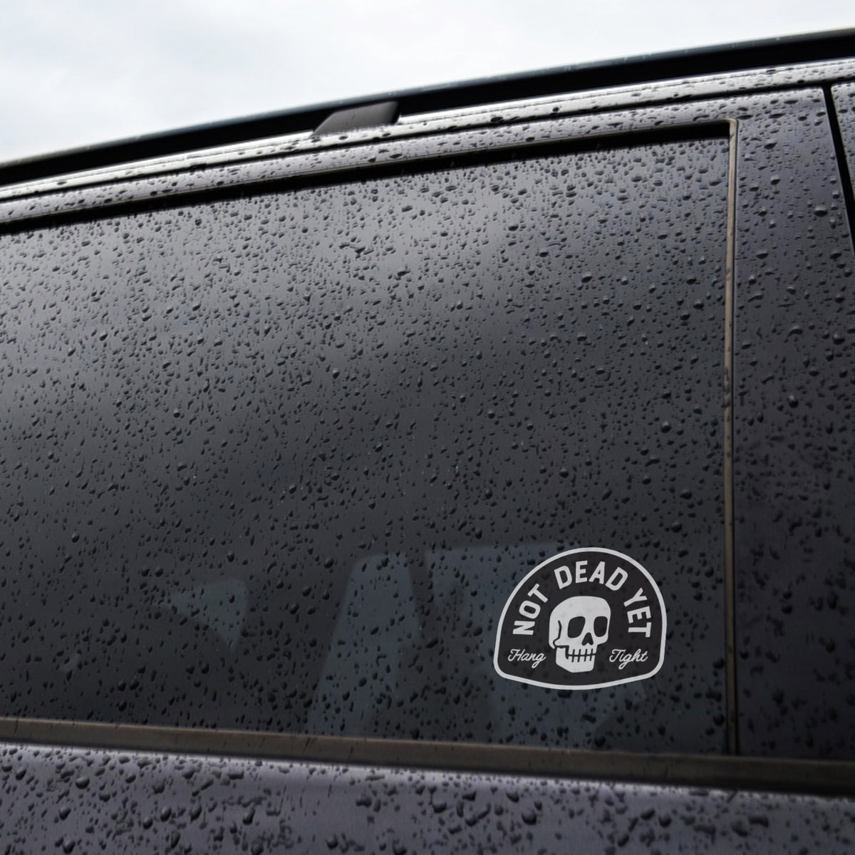 Raindrops cover a dark car window where the Not Dead Yet sticker sits near the lower corner.