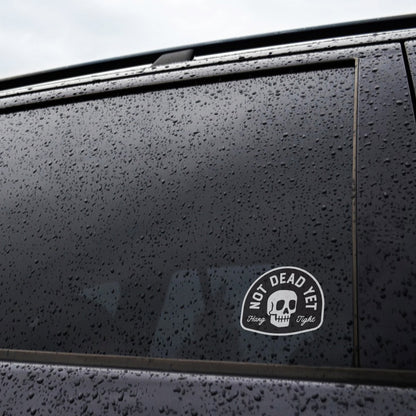 Raindrops cover a dark car window where the Not Dead Yet sticker sits near the lower corner.