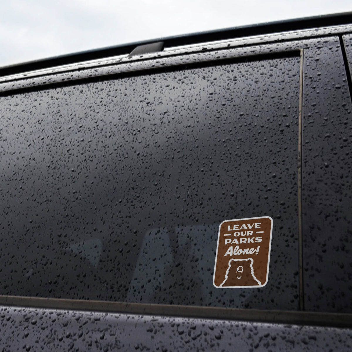 Rain beads across a car window where the Leave Our Parks Alone sticker sits near the bottom corner.