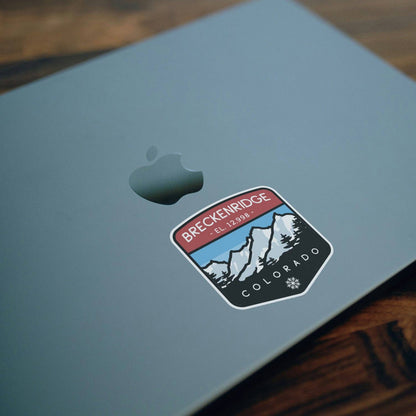 Stuck below the logo on a pale grey laptop lid, the Breckenridge Colorado sticker rests on dark wood.
