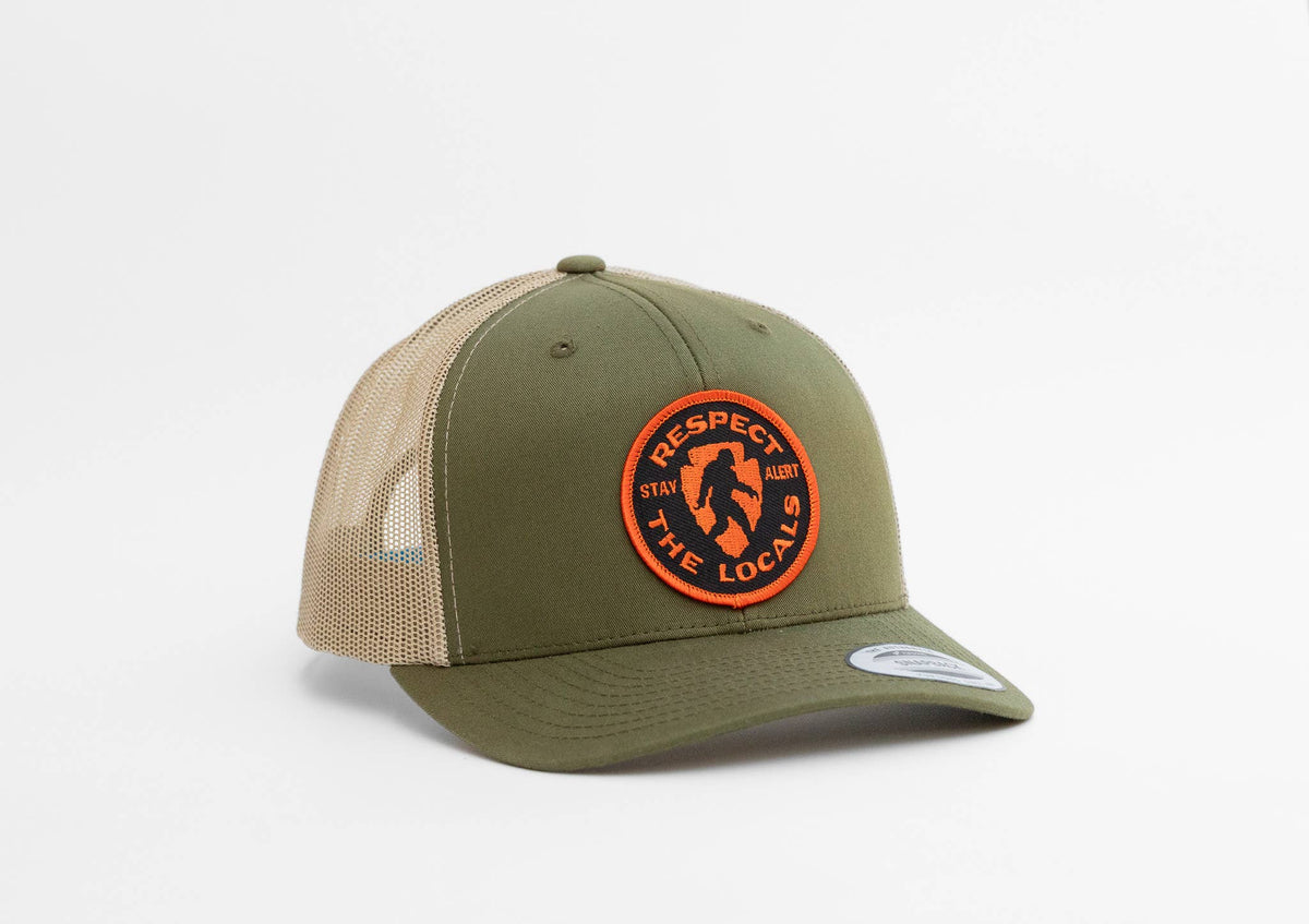 Olive and tan Bigfoot Respect The Locals trucker hat angled to show its round orange patch on the front panel.