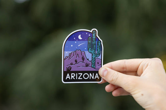 A hand holds the Arizona Desert Night Sky sticker, saguaro and purple buttes lit by stars, against green blur.