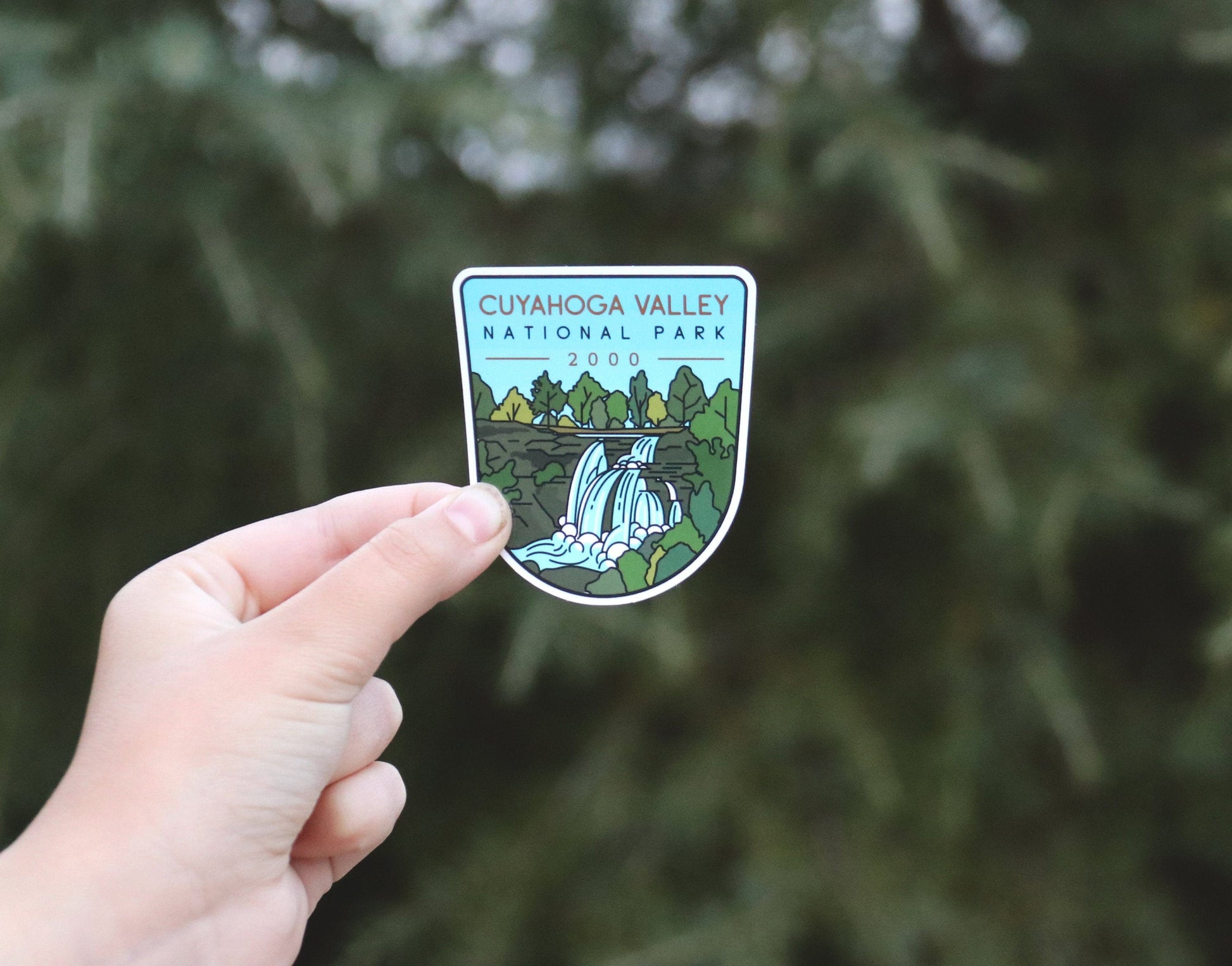 Held further back against blurred evergreen branches, the Cuyahoga Valley National Park sticker shows its full shield shape.
