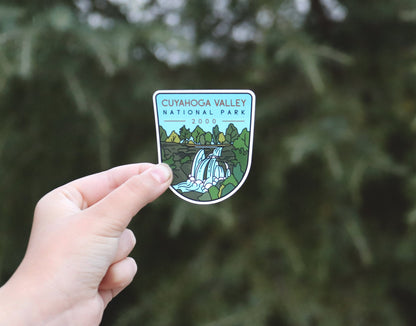 Held further back against blurred evergreen branches, the Cuyahoga Valley National Park sticker shows its full shield shape.