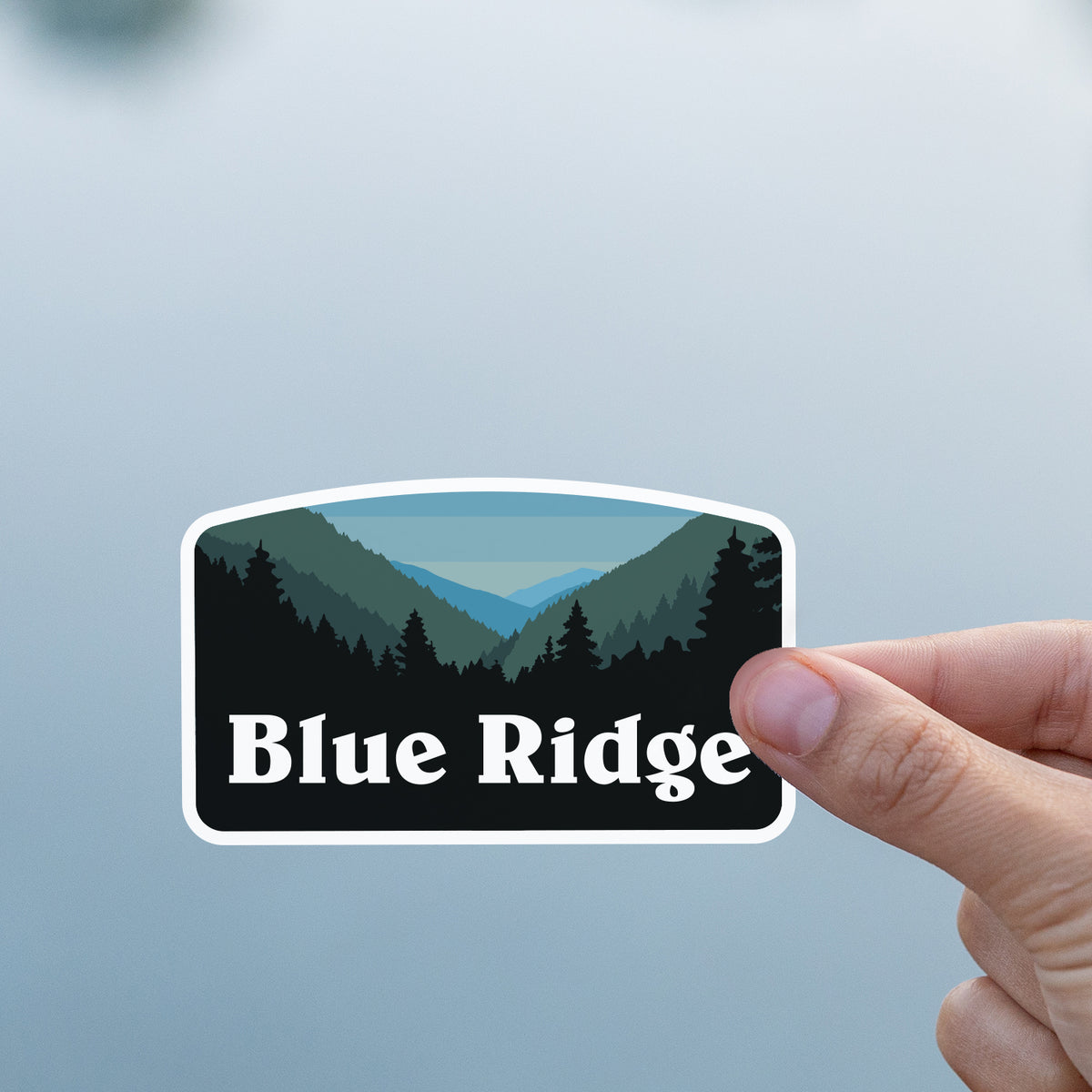 A thumb and finger hold the Blue Ridge sticker against a soft gray lake backdrop.