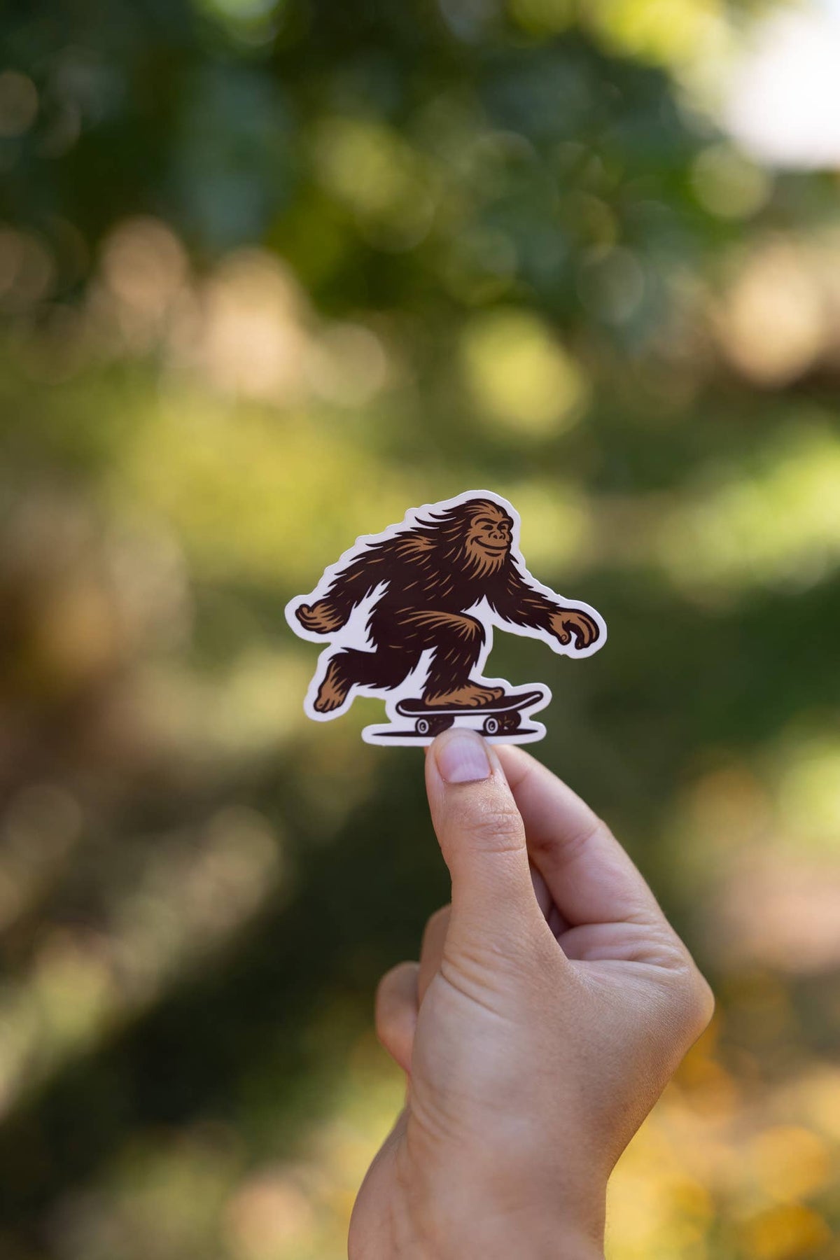 Held vertically toward a sunlit tree, the Later Skater Bigfoot sticker catches soft afternoon light.