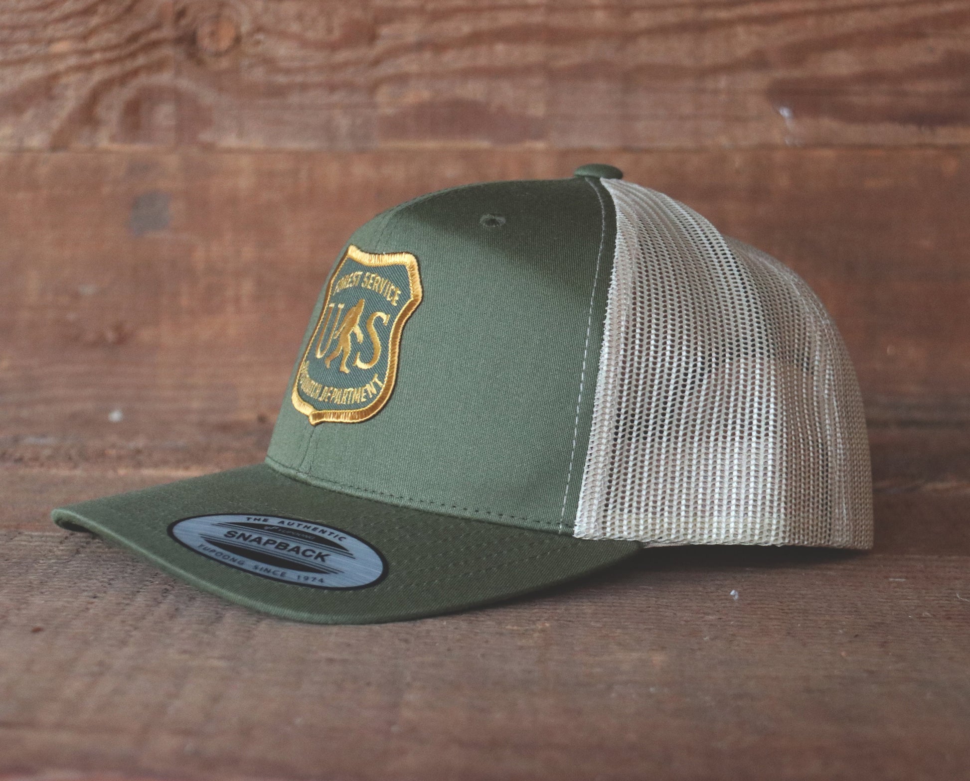 An olive and tan trucker hat carries the US Sasquatch Department patch on the front panel against barn wood.