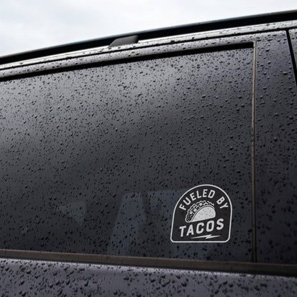Applied inside a rain beaded car window, the Fueled By Tacos sticker reads clearly on dark grey paint.