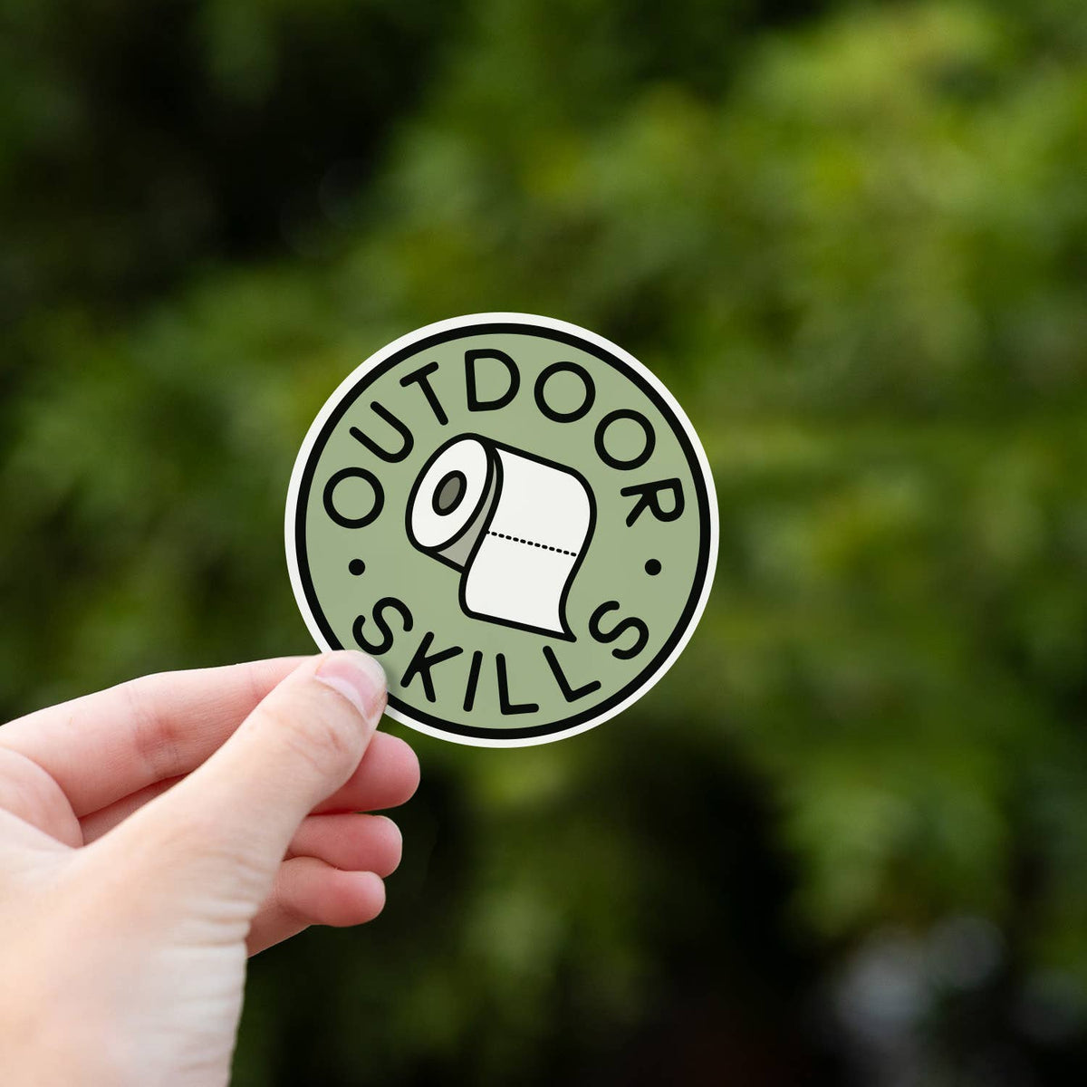 A hand raises the Outdoor Skills sticker toward the camera with green foliage falling out of focus.