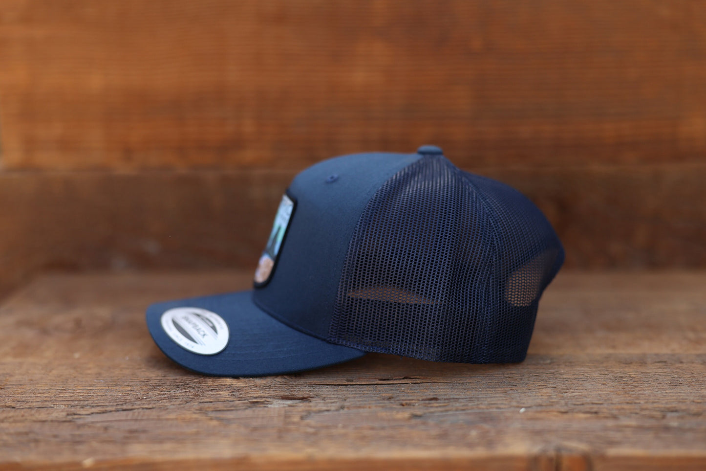 Side profile on rough wood, the navy Olympic National Park trucker hat shows mesh back and a curved brim.
