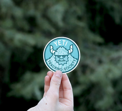 Backed by blurred evergreen branches, the Yeti Tracking Society sticker is pinched at arm's length.